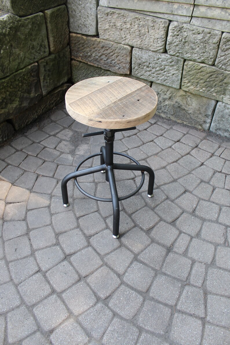 K&ouml;nnte beinhalten: Ein schwarzer Drehhocker aus Metall mit einem runden Holz-Sitz. Der Sitz ist aus recyceltem Holz und hat einen rustikalen Look. Der Hocker steht auf einer grauen Steinplatte.