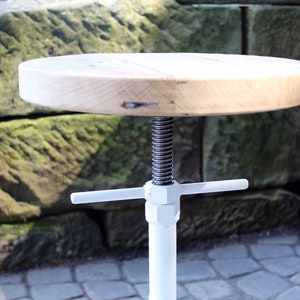 May include: White adjustable stool with a round wooden seat and a metal base. The base has a white finish and a screw mechanism for adjusting the height.