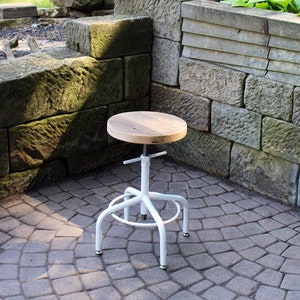 May include: White metal adjustable stool with a round wooden seat. The stool is sitting on a grey brick patio.