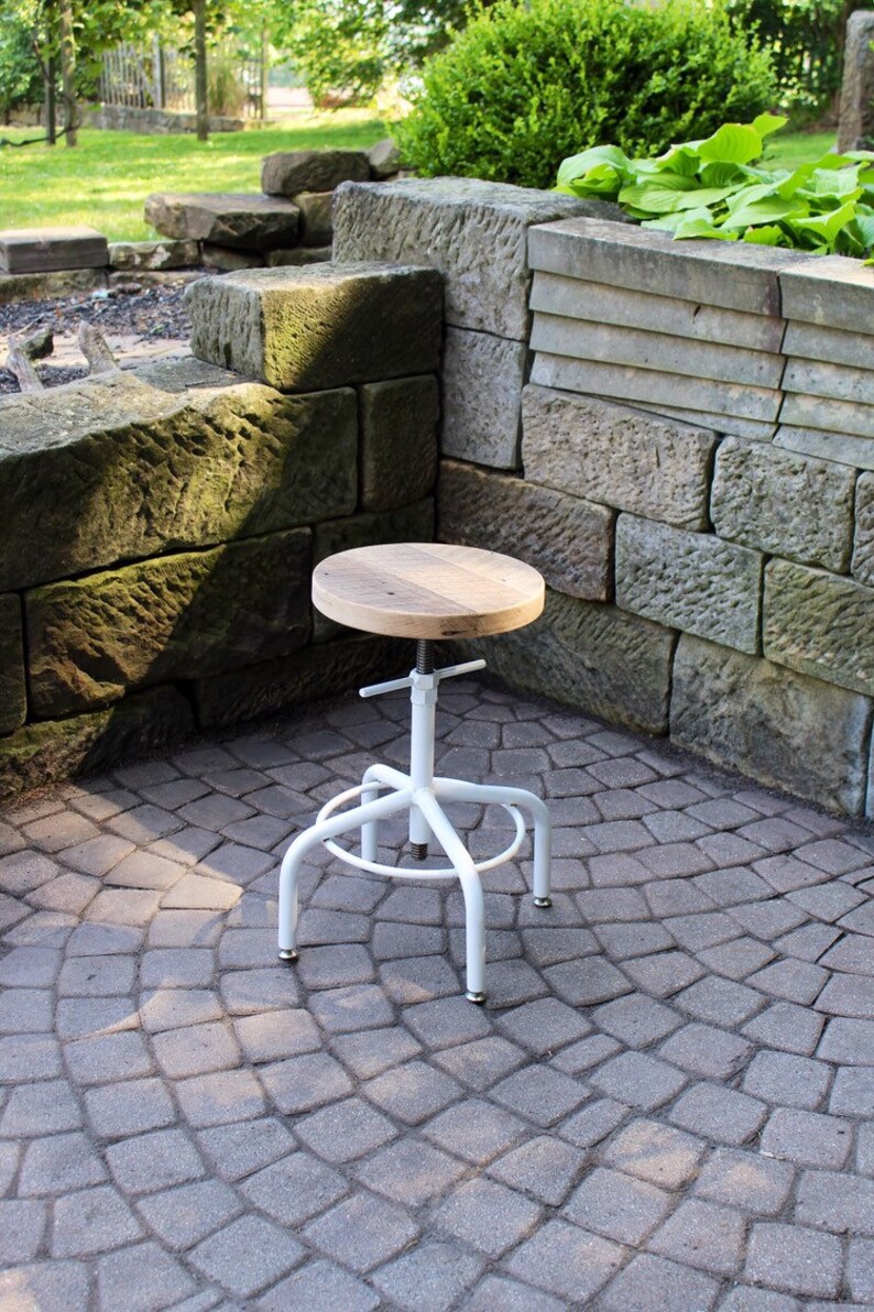 May include: White metal adjustable stool with a round wooden seat. The stool is sitting on a grey brick patio.