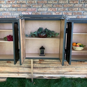 May include: A rustic wooden cabinet with three doors, each with a metal frame and hinges. The doors are open, revealing shelves inside. The cabinet is sitting on a stack of wooden planks.