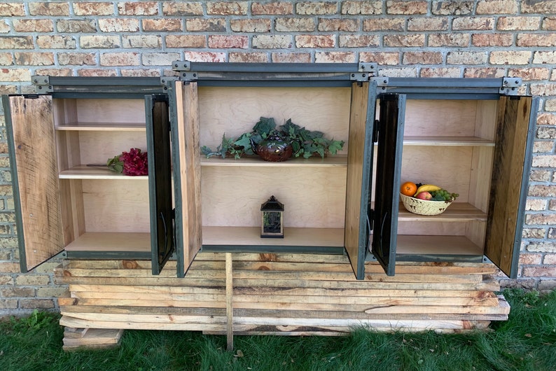 May include: A rustic wooden cabinet with three doors, each with a metal frame and hinges. The doors are open, revealing shelves inside. The cabinet is sitting on a stack of wooden planks.