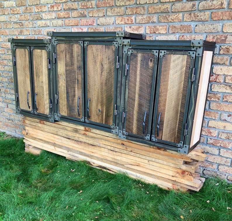May include: Three wooden cabinet doors with metal accents and hinges. The doors are painted a dark green colour and have a rustic wood grain. The cabinets are mounted on a wall and have a wooden shelf underneath.