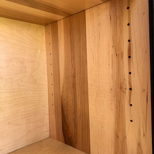 May include: Close-up of the interior of a wooden cabinet with a light wood finish. The cabinet has a shelf and several rows of evenly spaced holes drilled into the side panels.