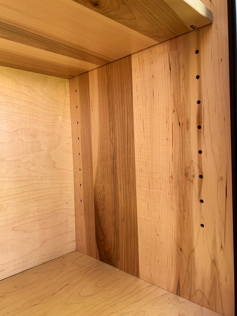 May include: Close-up of the interior of a wooden cabinet with a light wood finish. The cabinet has a shelf and several rows of evenly spaced holes drilled into the side panels.
