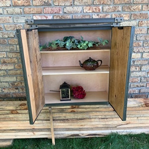 May include: A rustic wooden cabinet with two doors, a metal frame, and two shelves. The cabinet is open, revealing a teapot, a lantern, and a bouquet of flowers.