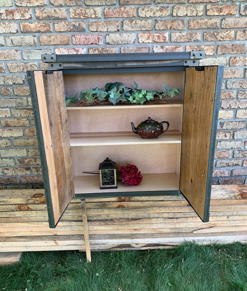 May include: A rustic wooden cabinet with two doors, a metal frame, and two shelves. The cabinet is open, revealing a teapot, a lantern, and a bouquet of flowers.