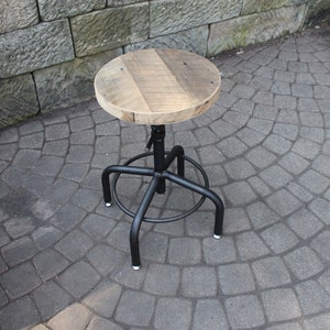 K&ouml;nnte beinhalten: Ein schwarzer Drehhocker aus Metall mit einer runden Holzfl&auml;che. Die Sitzfl&auml;che ist aus recyceltem Holz und hat eine nat&uuml;rliche Oberfl&auml;che. Der Hocker steht auf einer grauen Steinplatte.