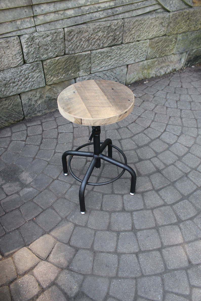 K&ouml;nnte beinhalten: Ein schwarzer Drehhocker aus Metall mit einer runden Holzfl&auml;che. Die Sitzfl&auml;che ist aus recyceltem Holz und hat eine nat&uuml;rliche Oberfl&auml;che. Der Hocker steht auf einer grauen Steinplatte.
