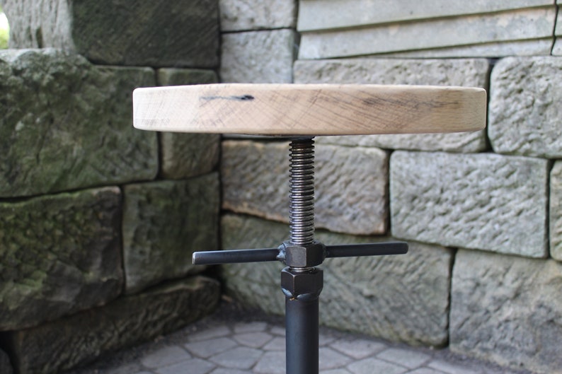 K&ouml;nnte beinhalten: Ein verstellbarer Hocker aus schwarzem Metall mit einer runden Holzfl&auml;che. Der Hocker verf&uuml;gt &uuml;ber einen Schraubmechanismus aus Metall zur H&ouml;henverstellung.