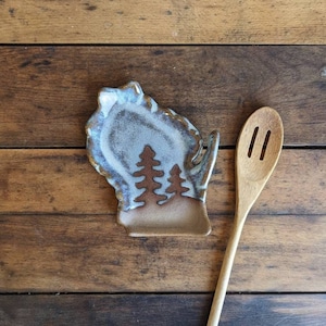 May include: A blue and brown ceramic spoon rest shaped like the state of Wisconsin with a silhouette of three pine trees. A wooden spoon is next to the rest.