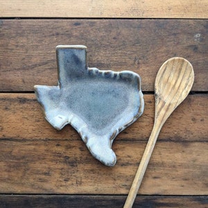 May include: A ceramic Texas-shaped spoon rest in a light blue and gray glaze. The rest is sitting on a wooden surface next to a wooden spoon.