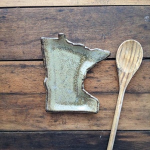 May include: A ceramic dish shaped like the state of Minnesota, with a light green glaze and brown accents. The dish is sitting on a wooden surface next to a wooden spoon.