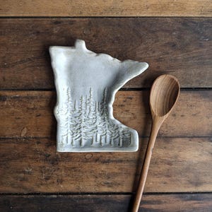May include: White ceramic spoon rest shaped like the state of Minnesota with a design of pine trees. A wooden spoon is next to the rest.