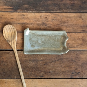 May include: A light brown wooden spoon and a light gray ceramic spoon rest with a slightly irregular shape. The spoon rest is sitting on a wooden surface.