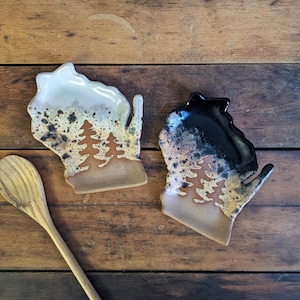 May include: Two ceramic spoon rests shaped like the state of Wisconsin. One is cream and brown with a tree design, the other is black and brown. A wooden spoon rests on a wooden surface.