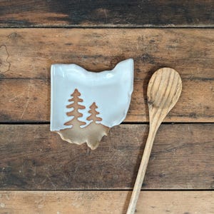 May include: A white ceramic spoon rest in the shape of Ohio, with two brown tree silhouettes. A wooden spoon rests beside it on a rustic wooden surface. The spoon rest is approximately 5 inches long.