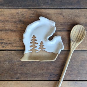 May include: A ceramic spoon rest in the shape of the state of Michigan, with a white glazed top and a tan base. Two stylized pine trees are cut out of the base. A wooden spoon rests on a wooden surface.