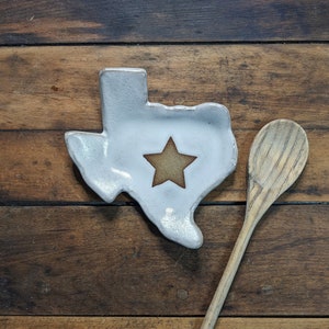 May include: White ceramic Texas-shaped spoon rest with a brown star in the center. The spoon rest is sitting on a wooden surface next to a wooden spoon.