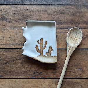 May include: A white ceramic spoon rest shaped like the state of Arizona, featuring a cactus design. The spoon rest is on a wooden surface, next to a wooden spoon. The spoon rest is approximately 5 inches long and 4 inches wide.
