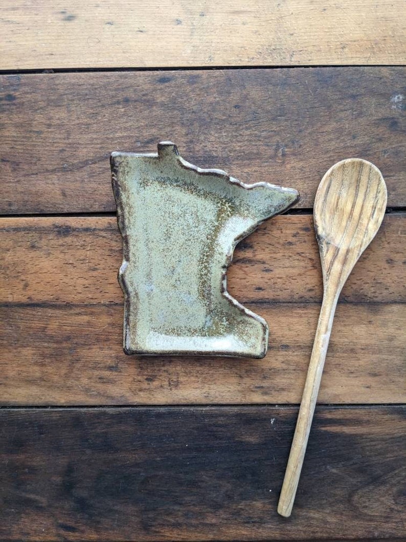 May include: A ceramic dish shaped like the state of Minnesota, with a light green glaze and brown accents. The dish is sitting on a wooden surface next to a wooden spoon.