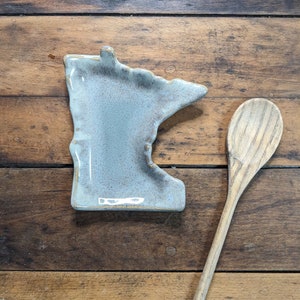 May include: A light blue ceramic dish shaped like the state of Minnesota. The dish is sitting on a wooden surface next to a wooden spoon.