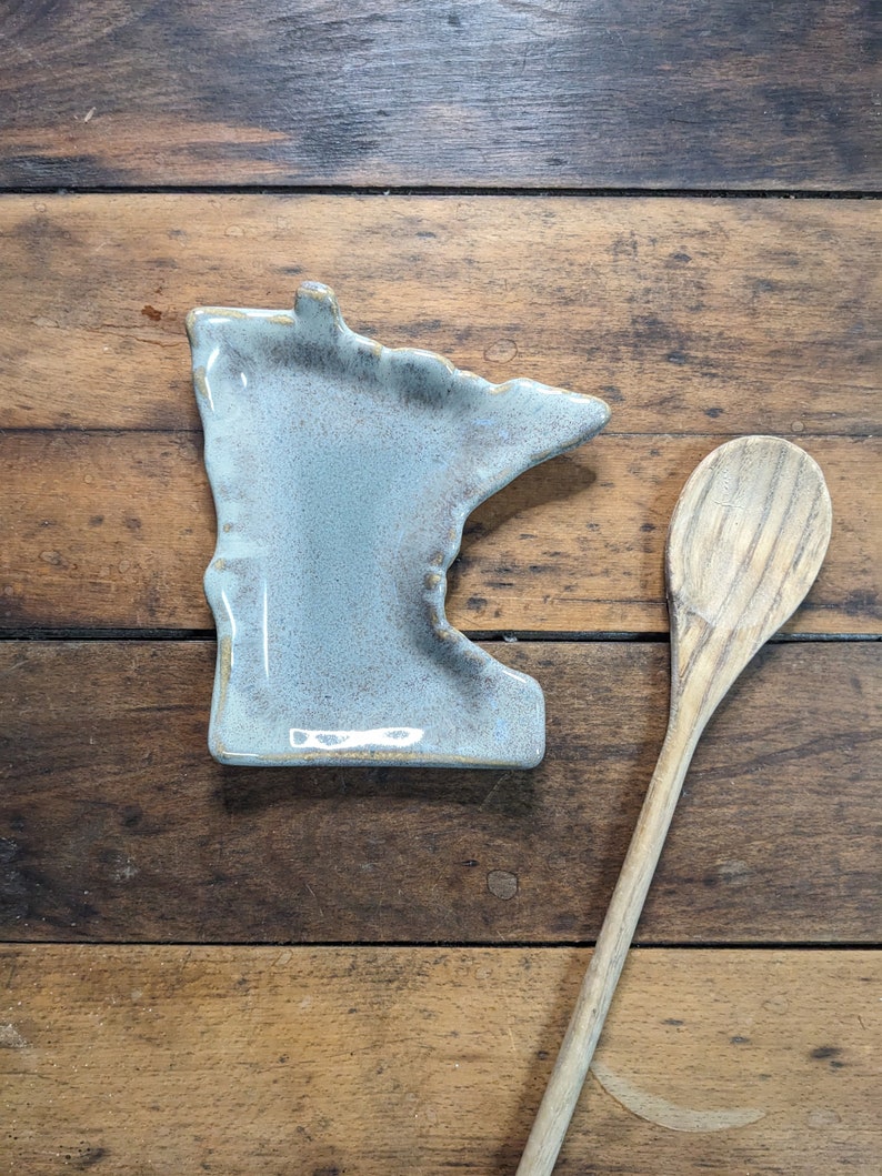 May include: A light blue ceramic dish shaped like the state of Minnesota. The dish is sitting on a wooden surface next to a wooden spoon.