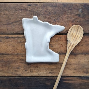 May include: A white ceramic spoon rest shaped like the state of Minnesota, next to a wooden spoon on a wooden surface.