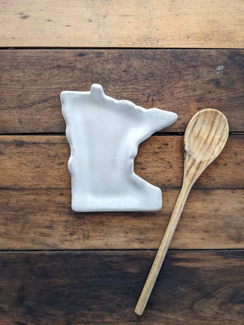 May include: A white ceramic spoon rest shaped like the state of Minnesota, next to a wooden spoon on a wooden surface.