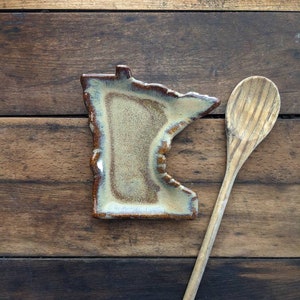 May include: A ceramic spoon rest shaped like the state of Minnesota, with a brown and beige glaze. A wooden spoon is placed next to the rest on a wooden surface.
