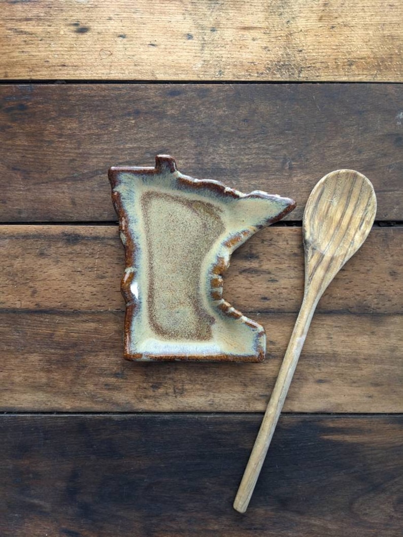 May include: A ceramic spoon rest shaped like the state of Minnesota, with a brown and beige glaze. A wooden spoon is placed next to the rest on a wooden surface.