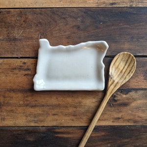 May include: A white ceramic dish shaped like the state of Oregon, sitting on a wooden surface with a wooden spoon beside it.