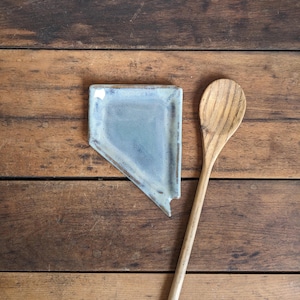 May include: A handmade, blue ceramic spoon rest in the shape of Nevada, next to a wooden spoon. The spoon rest has a rectangular shape with a cutout on one side. The wooden spoon is light brown and has a long handle.
