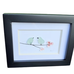 May include: A framed artwork featuring two light blue birds perched on a branch with pink flowers. The artwork is set against a white background and has a black frame.