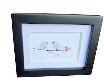 May include: A framed artwork featuring two light blue birds perched on a branch with pink flowers. The artwork is set against a white background and has a black frame.