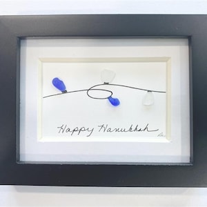 May include: Framed artwork with a black frame, featuring a white background with a line drawing of lights. Two blue and two white glass pieces are attached to the line. The words "Happy Hanukkah" are written in black script.