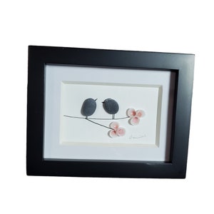 May include: Framed pebble art depicting two gray birds perched on a branch with pink flowers. The artwork is signed "Danielle".