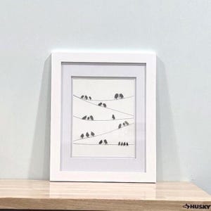 May include: A framed art print featuring a minimalist illustration of birds perched on power lines. The artwork is in grayscale, with a white frame and matting. The birds are depicted as simple black shapes.