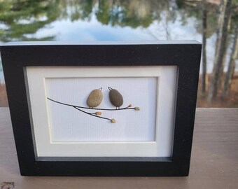 May include: A framed artwork featuring two small birds made of stones perched on a branch with small brown leaves. The artwork is on a white background and has a black frame.