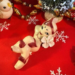 May include: A ceramic figurine of a reclining figure with a cream-colored body, maroon painted accents on the limbs and head, and a small white flower detail near the face. The figure has stylized facial features and is posed in a relaxed position.