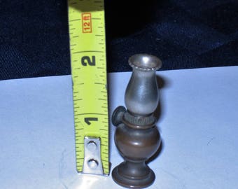 May include: A miniature oil lamp with a silver-colored glass chimney and a bronze-colored base. The lamp is about 2 inches tall and is sitting on a white surface.