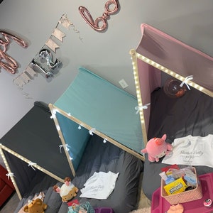 May include: Three teepees set up for a slumber party. The teepees are made of fabric and have wooden frames. The teepees are decorated with string lights and have pillows and blankets inside. The teepees are set up in a room with a gray wall and a red couch in the background. The teepees are decorated with a banner that says "Happy 12th Birthday".