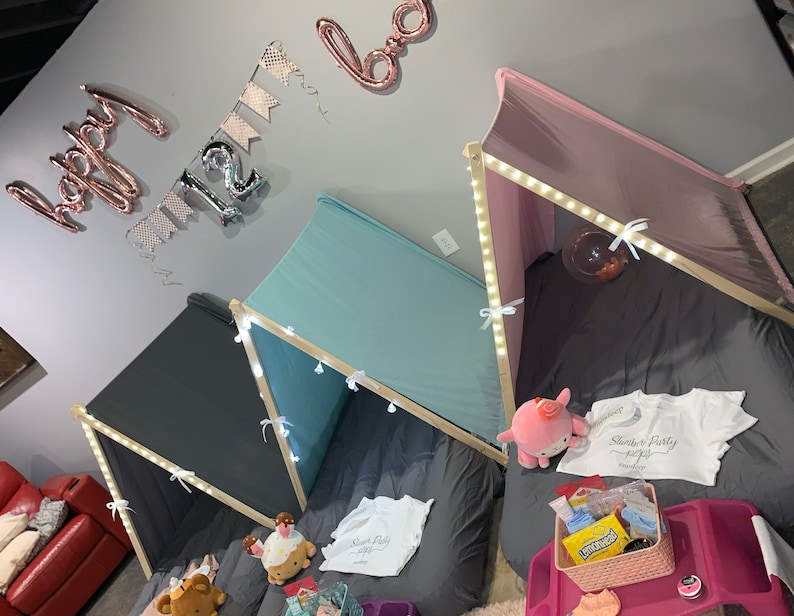 May include: Three teepees set up for a slumber party. The teepees are made of fabric and have wooden frames. The teepees are decorated with string lights and have pillows and blankets inside. The teepees are set up in a room with a gray wall and a red couch in the background. The teepees are decorated with a banner that says "Happy 12th Birthday".