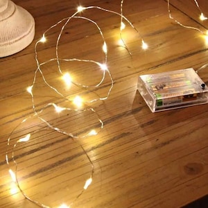 May include: A string of warm white LED fairy lights with a clear battery pack. The lights are wrapped around a wooden surface.