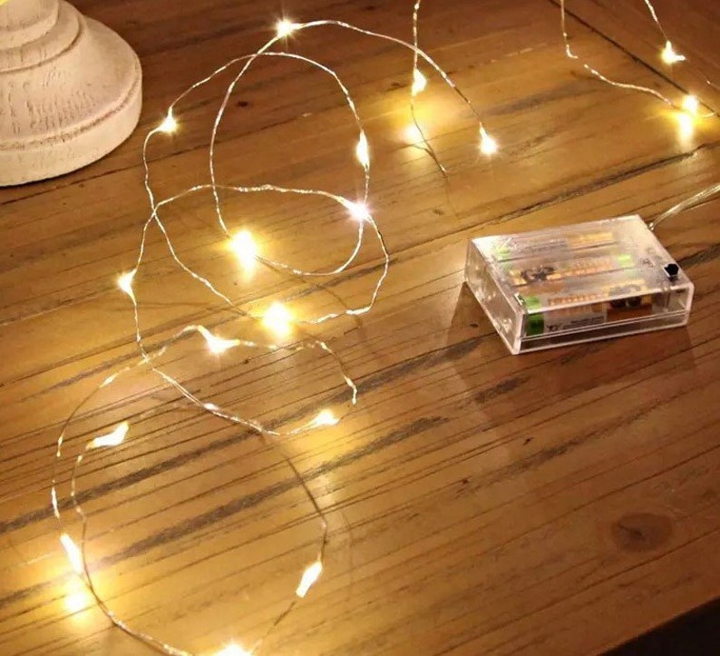 May include: A string of warm white LED fairy lights with a clear battery pack. The lights are wrapped around a wooden surface.