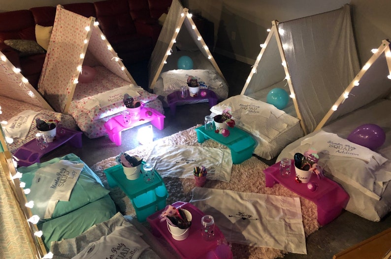 May include: A sleepover setup with multiple teepees decorated with fairy lights and personalized pillows. Each teepee has a small table with a bucket, cups, and snacks. The teepees are on a fluffy rug with a few balloons.