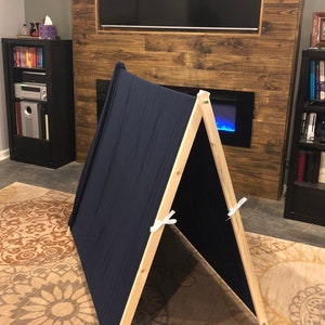 May include: A simple wooden A-frame tent with a dark blue fabric cover. The tent is set up on a patterned rug in a living room.