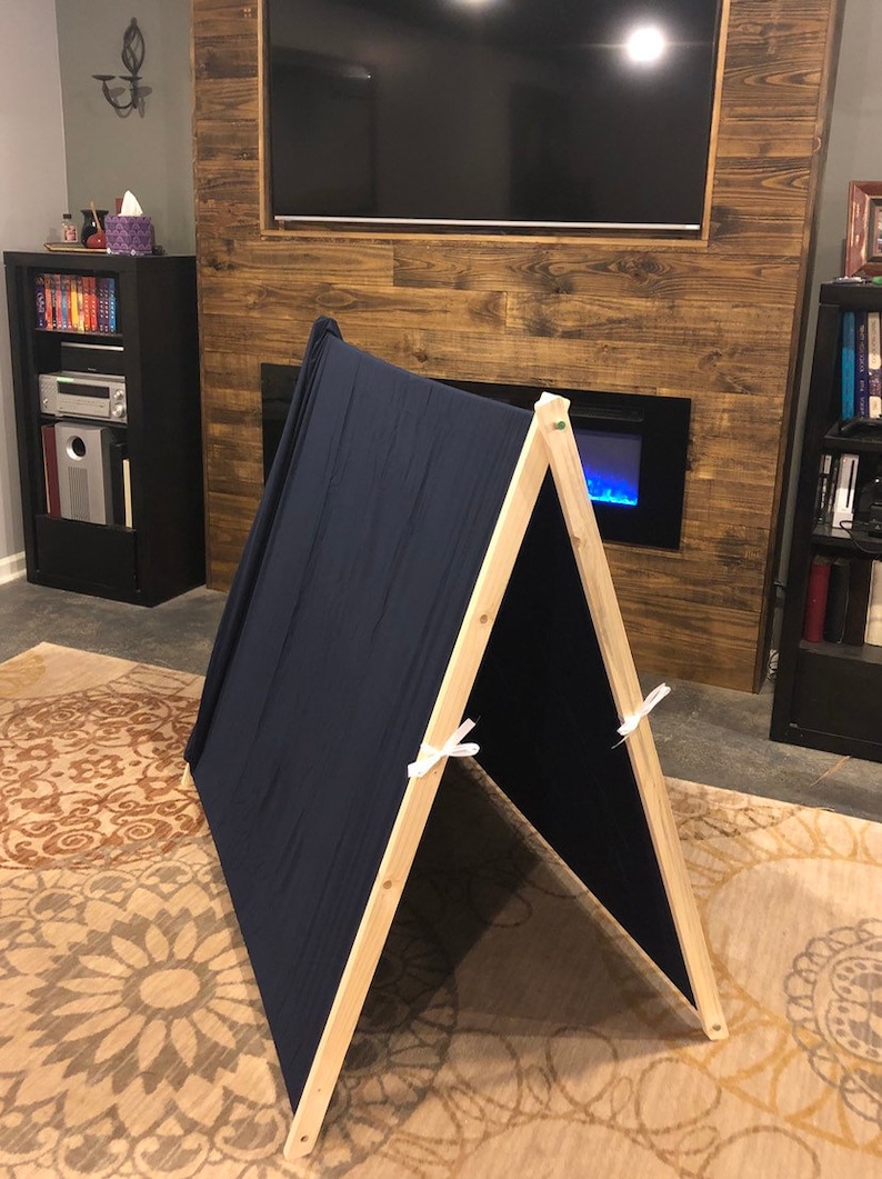 May include: A simple wooden A-frame tent with a dark blue fabric cover. The tent is set up on a patterned rug in a living room.