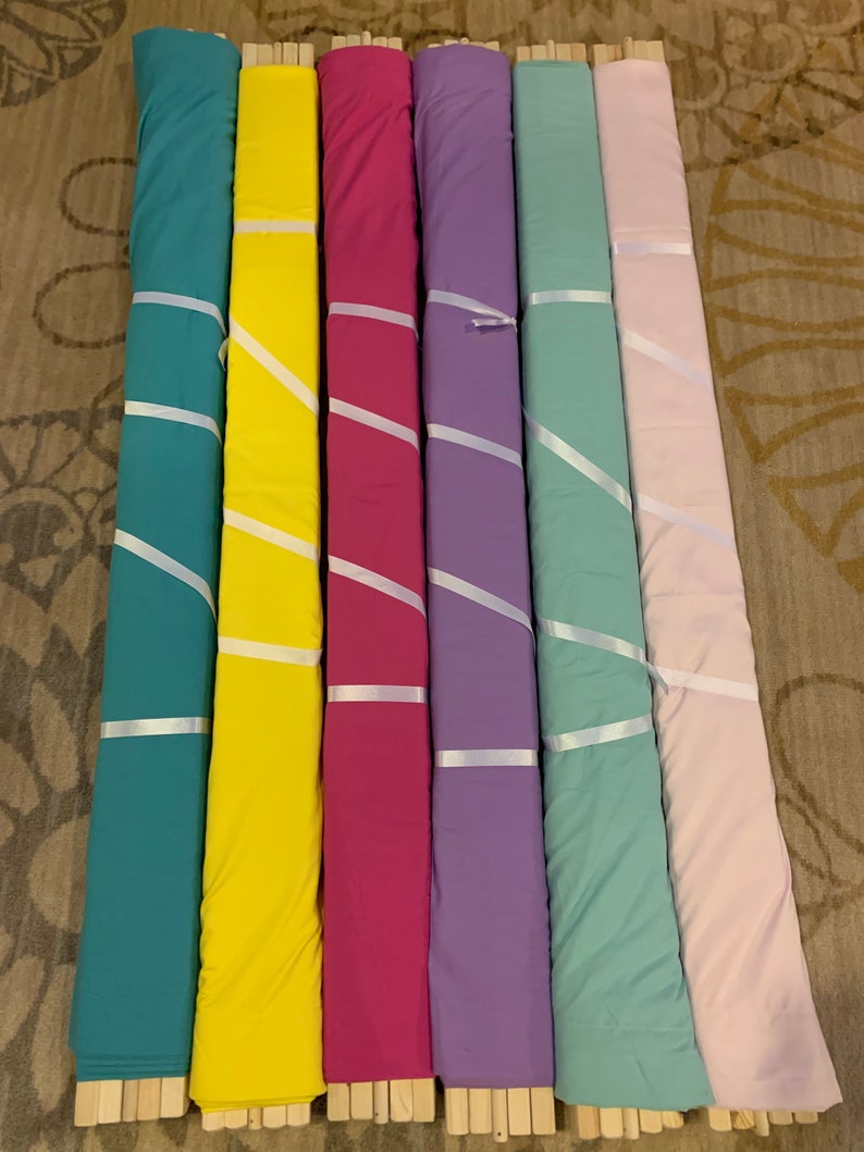 May include: Seven rolls of fabric in various colors, including teal, yellow, pink, purple, light green, and light pink. The fabric is rolled up and tied with white ribbon. The fabric is displayed on a wooden surface.