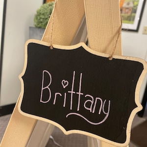 May include: A black chalkboard sign with a wooden frame hangs from a string. The sign says "Brittany" in pink cursive with a small pink heart.